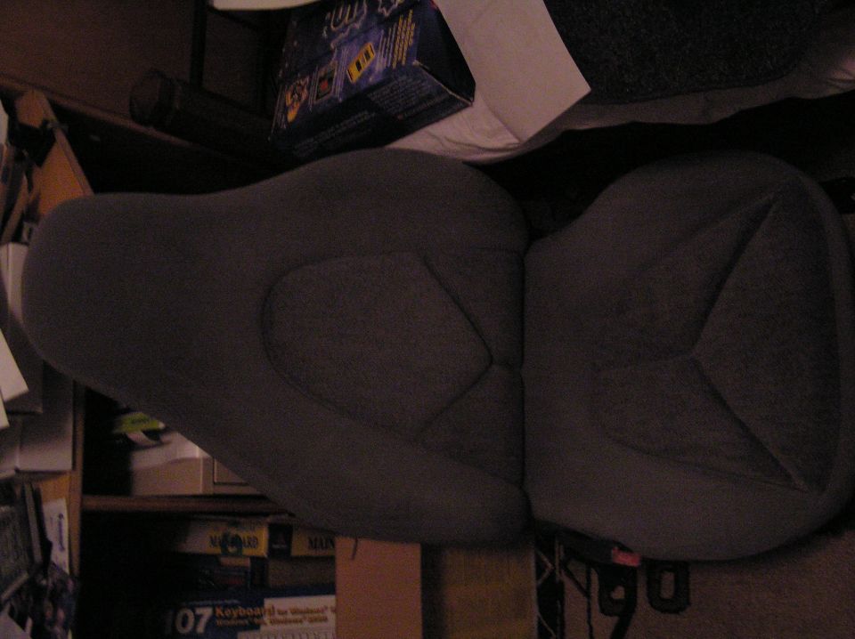 Two dark gray cloth car seats set out indoors with shelving and boxes behind