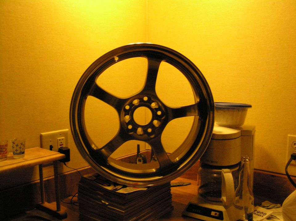 Gunmetal five-spoke alloy wheel standing on magazines beside a coffee maker, yellow wall