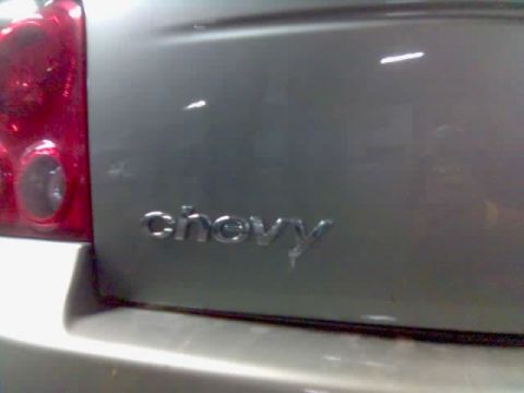 1985 Ford Bronco - Close-up of a 'chevy' emblem on a silver car's trunk lid, with a red taillight visible on the left.