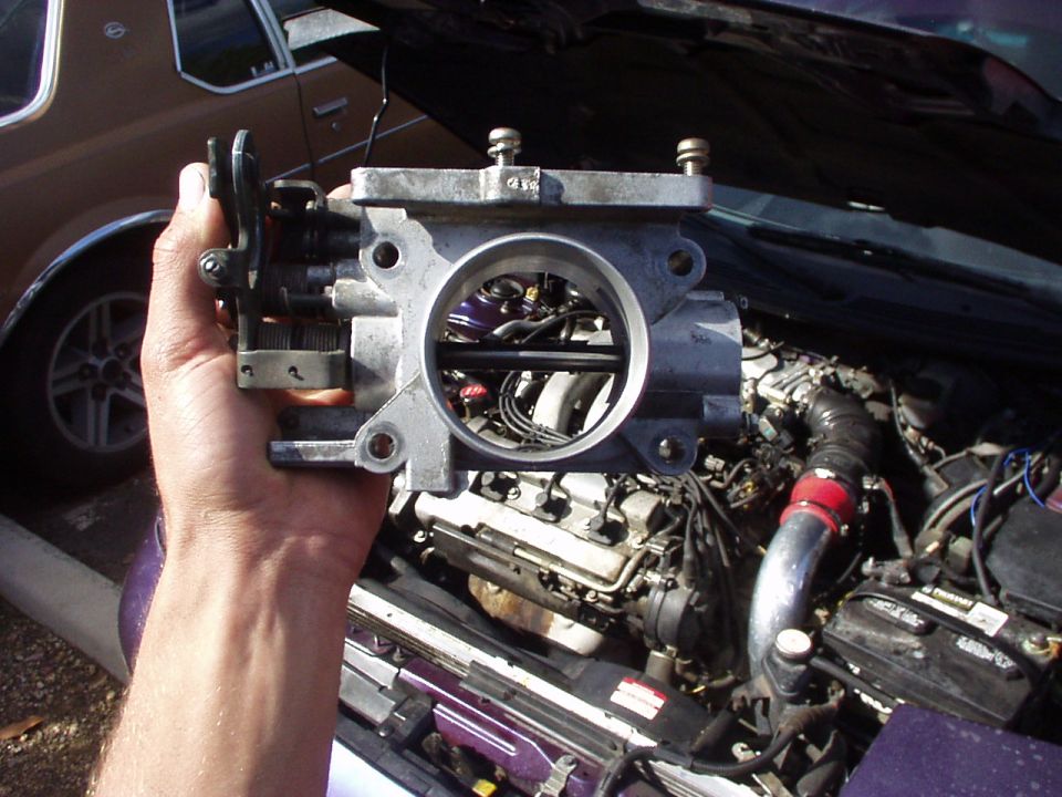Hand holding a removed throttle body over an open engine bay with intake and engine visible