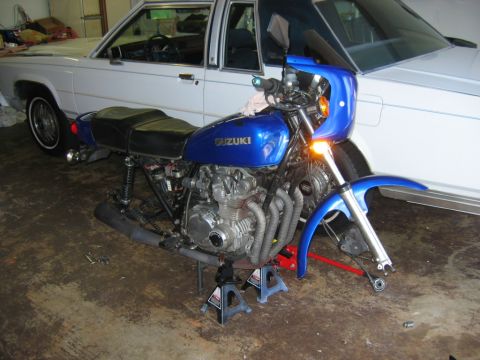 1985 Ford Crown Victoria LTD - A blue Suzuki motorcycle is parked in a garage in front of a white sedan.