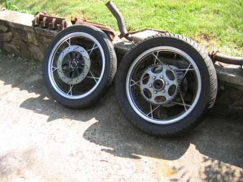 1985 Ford Crown Victoria LTD - Two motorcycle wheels, one a disc brake and the other a sprocket, lie next to exhaust pipes on a paved surface.