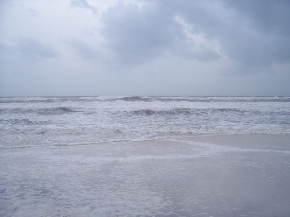 Ocean waves crashing on a sandy beach under a cloudy sky. A "SUPERMOTORS" watermark is visible in the lower right corner