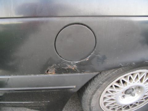 1990 Ford Taurus - Close-up of a car's rear quarter panel showing rust damage near the fuel filler door and a portion of the wheel.
