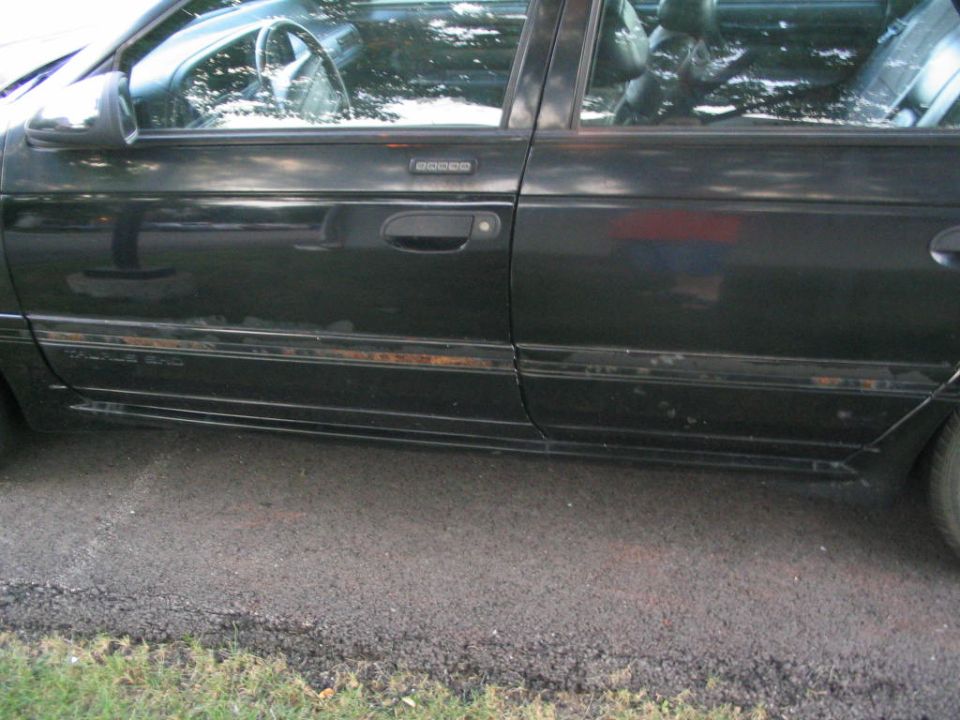 Side view of a black sedan with rust spots along the bottom trim. Interior visible through the windows