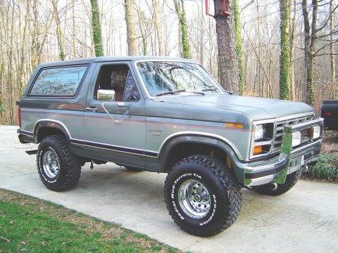 1986 Ford Bronco - A two-tone gray 1980s Ford Bronco full-size SUV shown from the front three-quarter angle, lifted on chrome wheels with BFGoodrich Mud-Terra…