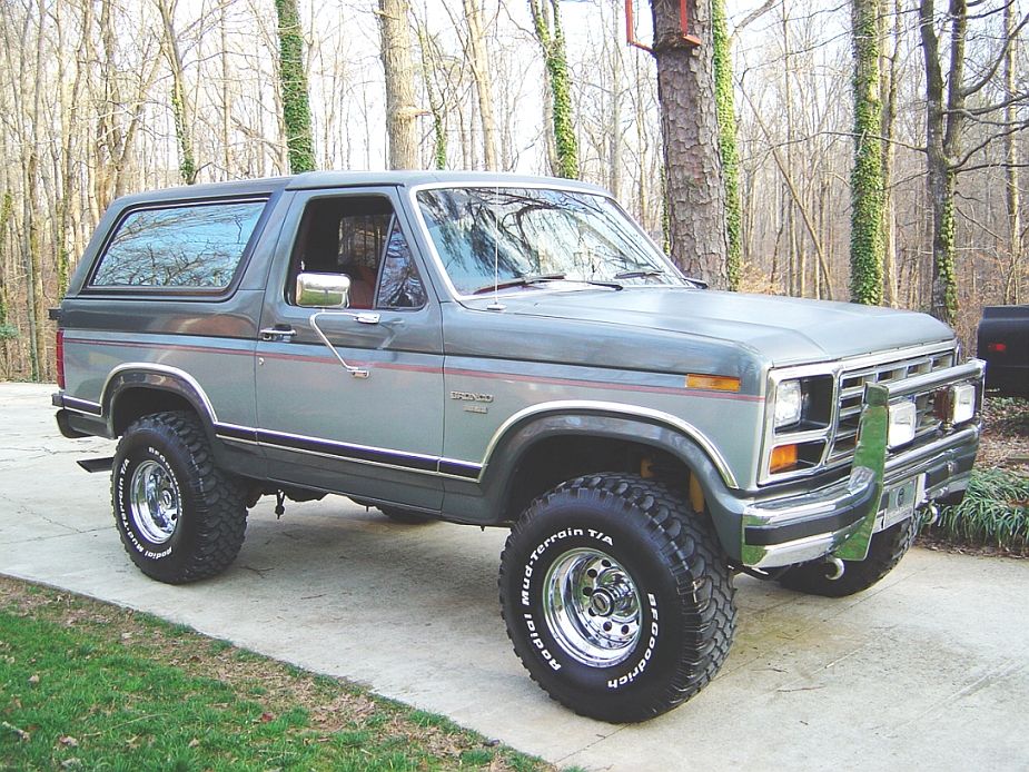 Gray lifted Ford Bronco SUV on chrome wheels, front 3/4 view, parked by woods.