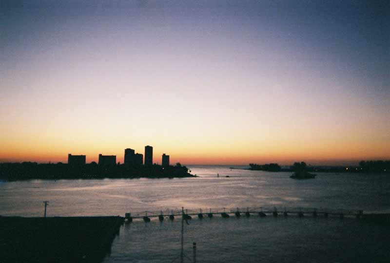 City skyline silhouetted against a sunset over the water, with a bridge in the foreground