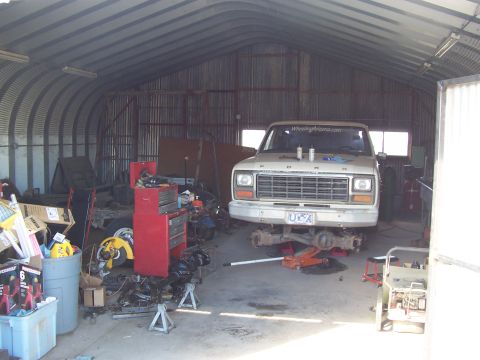 1981 Ford F-150 - A white Ford pickup truck sits front-forward inside a metal-arched workshop, raised on a red floor jack with its front axle removed and res…