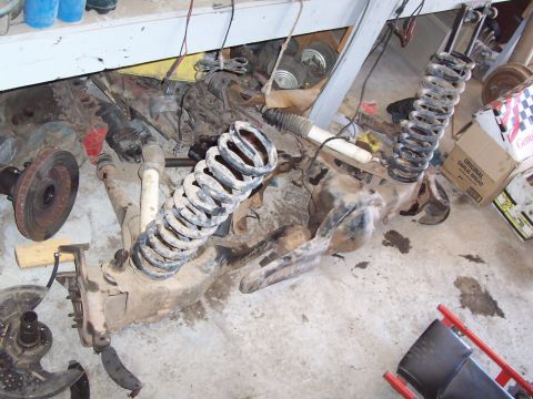 1981 Ford F-150 - Overhead view of a truck's front suspension and axle assembly disassembled on a garage floor, showing coil springs, shock absorbers, brake…