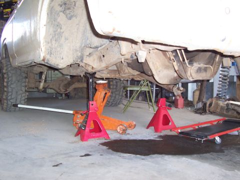 1981 Ford F-150 - Underside view of a white pickup truck raised on a floor jack and red jack stands in a garage, showing the frame rail, leaf/coil suspension…