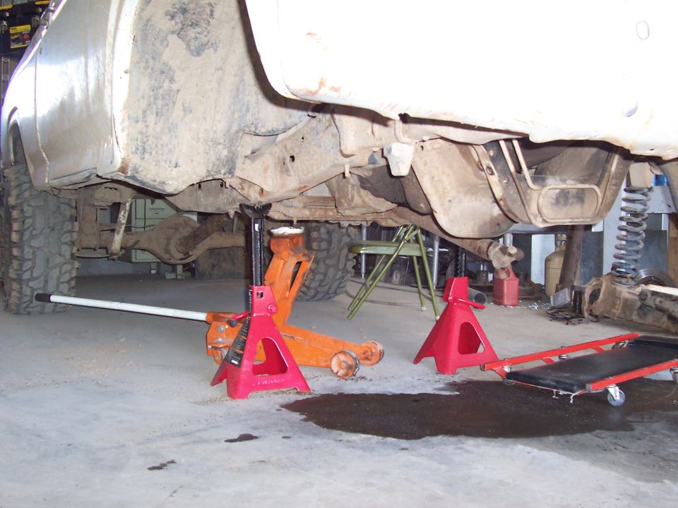 White pickup raised on floor jack and red jack stands showing undercarriage in garage