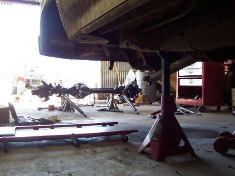 1981 Ford F-150 - Underside view of a truck raised on a red jack stand inside a garage, showing the frame rails, driveshaft, and a removed axle assembly rest…