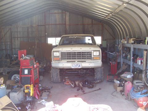 1981 Ford F-150 - Front view of a white Ford pickup truck raised on large off-road tires inside a metal-arched workshop, with the front axle/driveshaft remov…