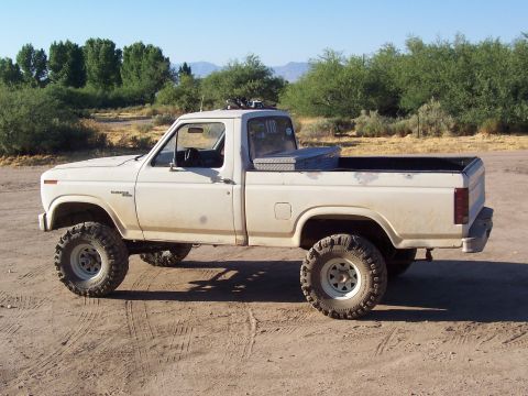 1981 Ford F-150 - This lifted pickup truck has a toolbox mounted in the bed and is parked on a dirt lot under a clear sky.