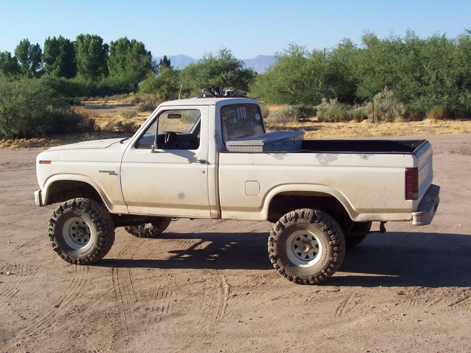 White pickup truck with large tires, lifted suspension, and a toolbox in the bed. It's parked on a dirt surface with trees and hills in the background