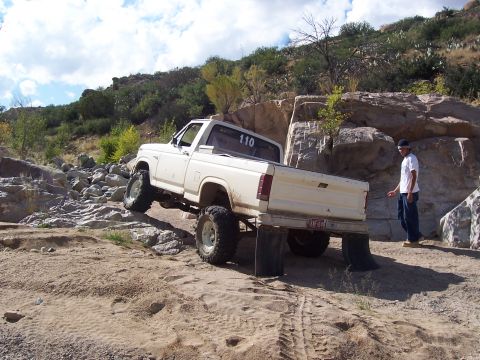 1981 Ford F-150 - A white pickup truck with oversized tires is shown off-roading on a rocky, sandy trail.