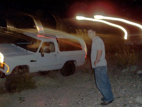 1981 Ford F-150 - A man stands beside a white SUV parked on a dirt lot at night, with light trails streaking across the image.