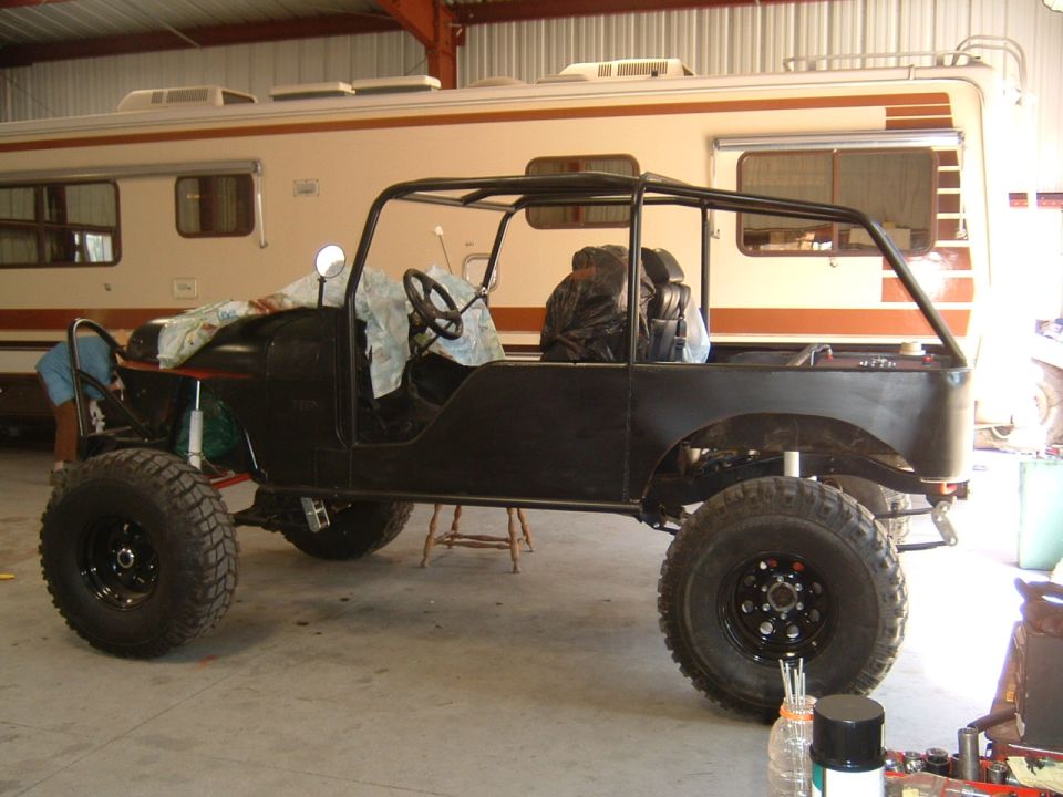 Black Jeep CJ with roll cage, lift, and big tires in a shop beside an RV