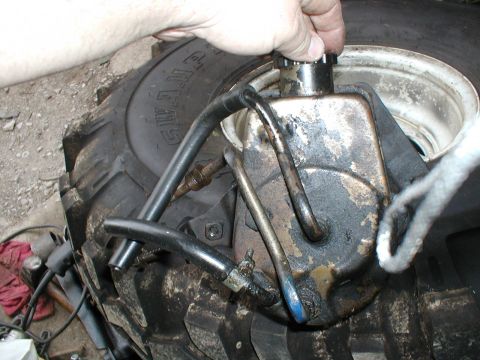 1979 Ford Bronco - A greasy power steering pump is shown with hoses connected, positioned next to a large tire. A hand is seen adjusting the pump's cap.