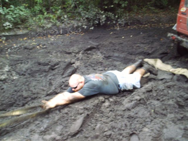 Man in dark t-shirt and shorts crawling through thick mud, reaching for a rope. A red vehicle is partially visible in the background