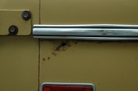 1972 Ford Bronco - Close-up of a yellow vehicle's side panel showing chrome trim, rust damage, and a portion of a red taillight.