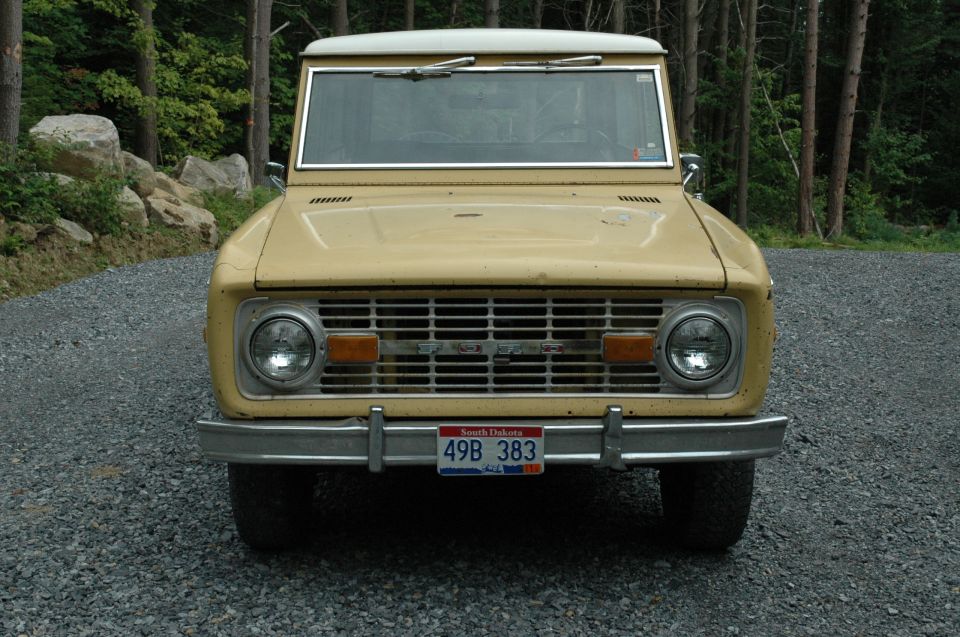 Front view of a tan Ford Bronco parked on a gravel driveway with trees in the background
