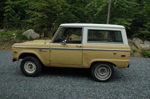 1972 Ford Bronco - Side profile of a tan and white 1972 Ford Bronco parked on a gravel surface with trees in the background.