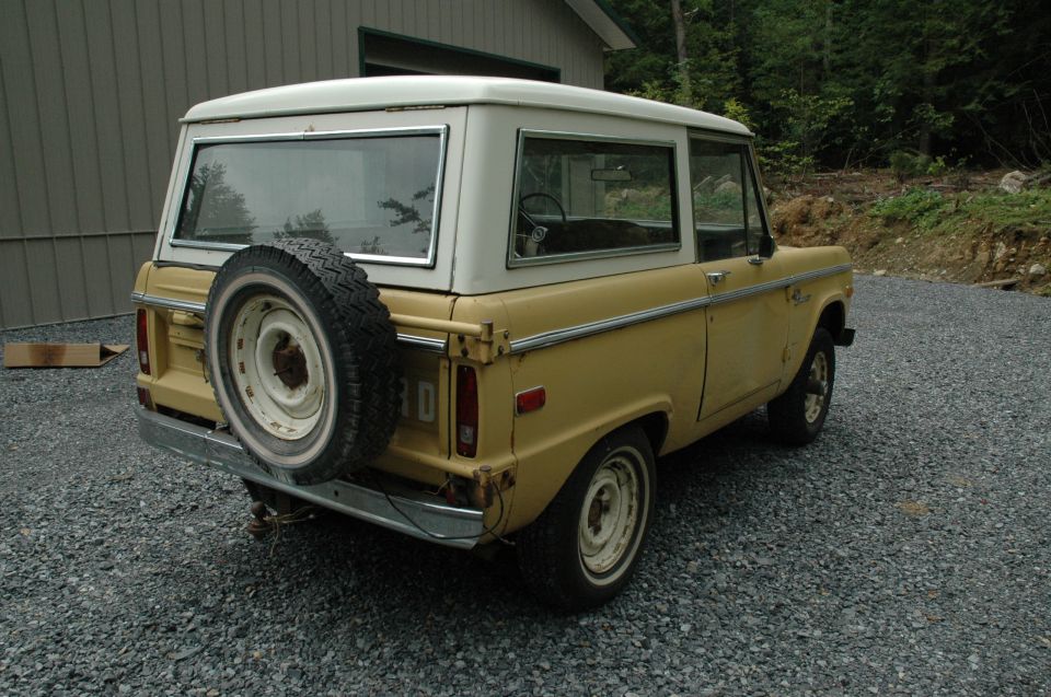 Tan early Ford Bronco with white hardtop and tailgate spare, rear three-quarter view on gravel