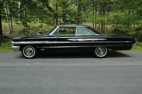 1964 Ford Galaxie - This black 1964 Ford Galaxie 500XL coupe is seen from the side, parked on a road with trees in the background.