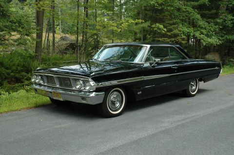 1964 Ford Galaxie - A black 1964 Ford Galaxie 500XL is parked on a road with a wooded area behind it.