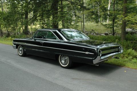 1964 Ford Galaxie - A black 1964 Ford Galaxie 500XL coupe is seen from a three-quarter front angle on a paved road beside a wooded area.