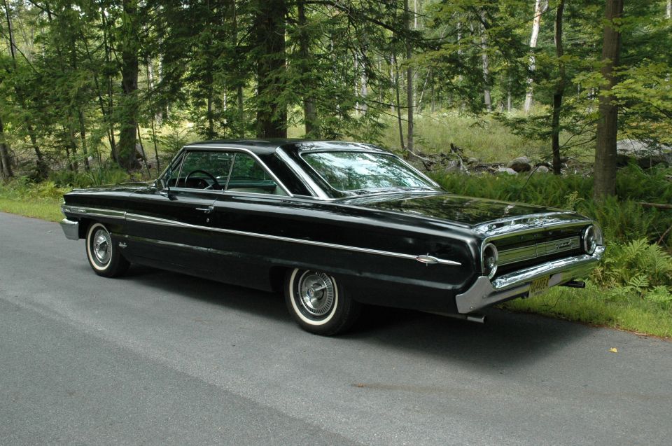 Black 1960s Ford Galaxie hardtop coupe, rear 3/4 view, parked by forest