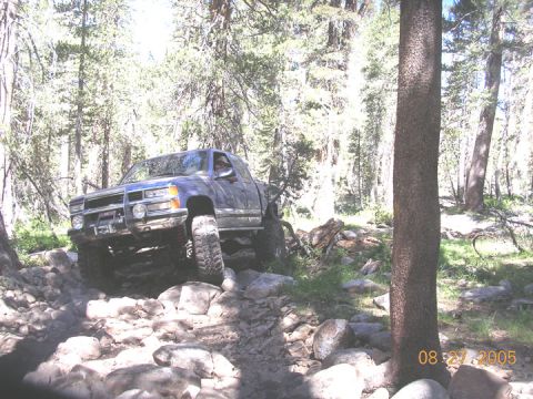 1994 Ford Bronco - This blue pickup truck is shown off-roading on a challenging rocky path through a wooded area.