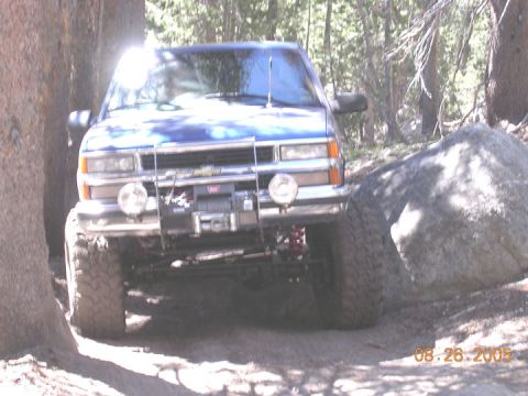1994 Ford Bronco - A blue Chevrolet pickup truck is seen off-roading on a rocky trail with trees and a large boulder.