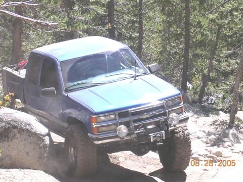 1994 Ford Bronco - Pickup truck driving over rocks on a dirt path with trees in the background.