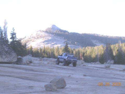 1994 Ford Bronco - A blue pickup truck navigates a rocky dirt road, with a mountain range and pine forest visible in the distance.