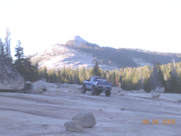 Blue pickup truck driving on a dirt road with mountains and trees in background