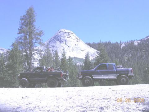 1994 Ford Bronco - Two off-road vehicles, a Ford Bronco and a Chevrolet pickup, are parked side-by-side on a dirt path with a large mountain and forest in the…