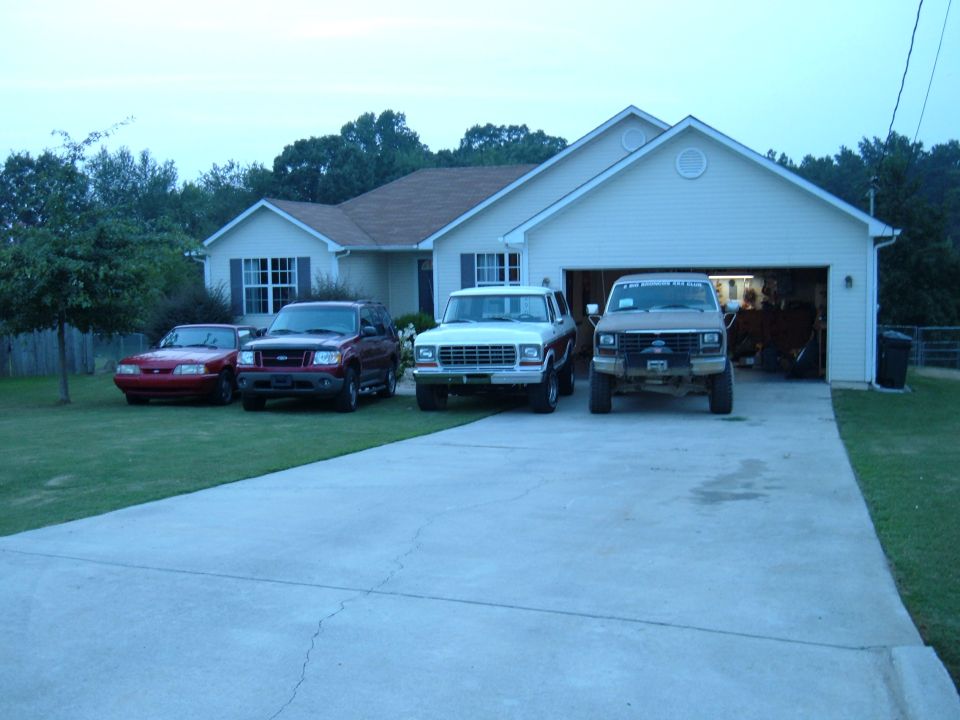 Red Mustang, maroon Explorer, white Ford pickup and brown lifted Bronco parked at a white house driveway