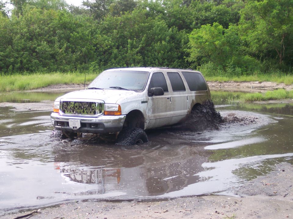 White lifted Ford Excursion SUV splashing through deep mud hole, front 3/4 view