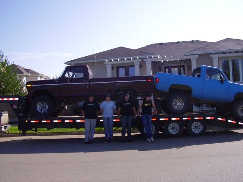 1979 Ford F-250 - Two lifted pickup trucks are being transported on a trailer, with four people posing in front of them.