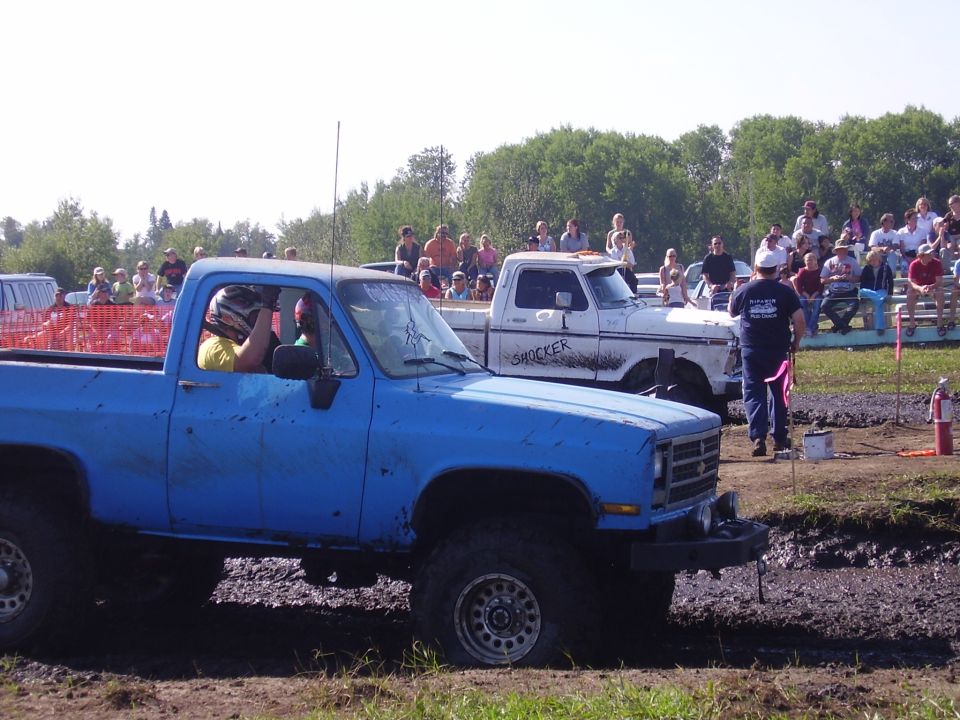 Blue pickup truck with mud splatter, driver wearing helmet, in a mud pit with spectators