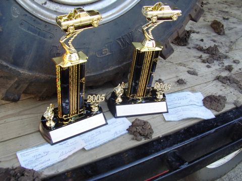 1979 Ford F-250 - Two trophies, dated 2005, are displayed on a trailer next to a large tire, with scattered dirt and papers.