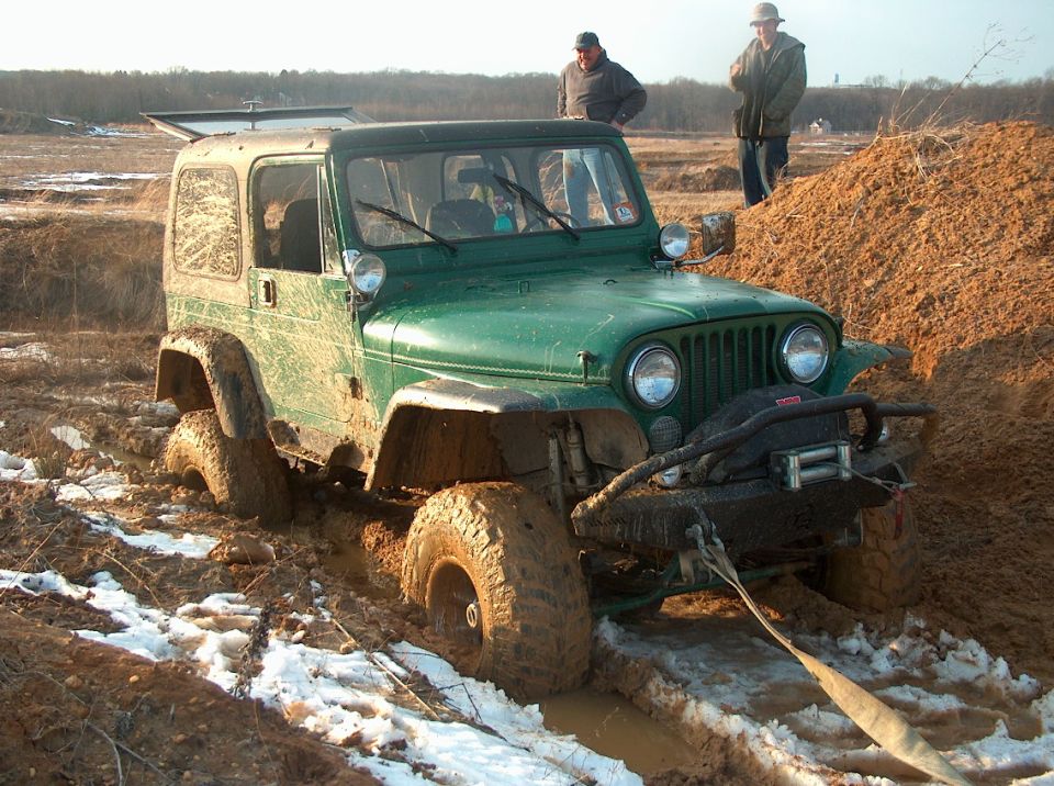 Green hardtop Jeep with big mud tires and winch stuck in muddy ruts with snow, recovery strap attached