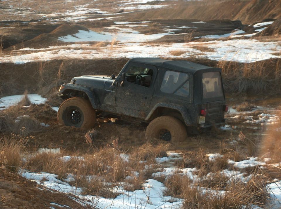 Mud-covered black Jeep Wrangler stuck in muddy terrain with patches of snow and dry grass