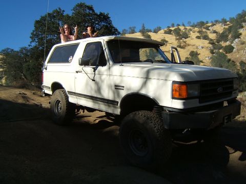 1988 Ford Bronco - A white Ford Bronco with two people on its roof drives on a dirt road with a hilly, tree-covered background.