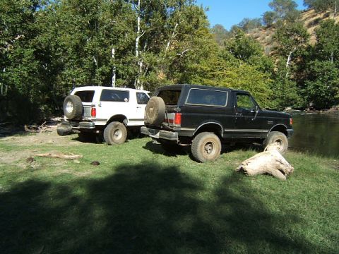 1988 Ford Bronco - Two Ford Broncos, a black and a white model, are parked on a grassy bank next to a flowing river.