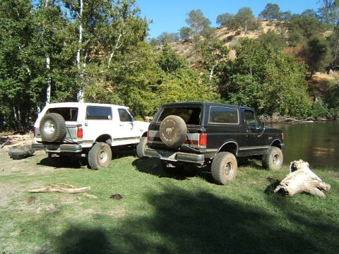 1988 Ford Bronco - Two Ford Broncos are parked near a river on a sunny day.