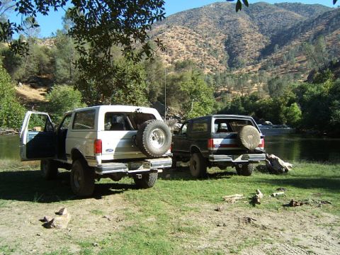 1988 Ford Bronco - Two Ford Broncos are parked near a river on a sunny day with hills in the background.